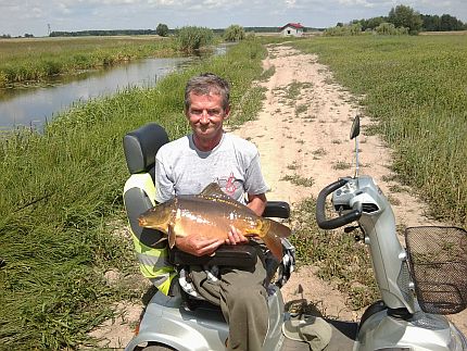 Mężczyzna na skuterze inwalidzkim trzyma dużą rybę, w tle pole i rzeczka