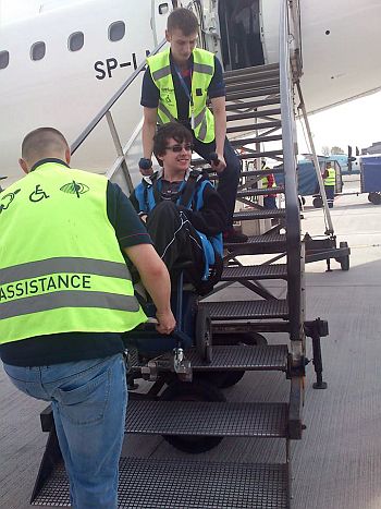 Dwóch asystentów w odblaskowych kamizelkach wnosi po schodkach do samolotu młodego mężczyznę na wózku