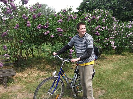 Młody mężczyzna stoi trzymając rower na tle kwitnącego bzu