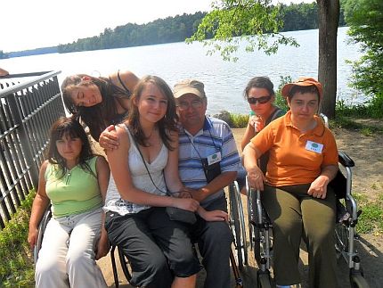 Grupa osób w różnym wieku pozuje do zdjęcia na tle jeziora, część osób jest na wózkach, na pierwszym planie młoda kobieta siedzi na kolanach mężczyzny