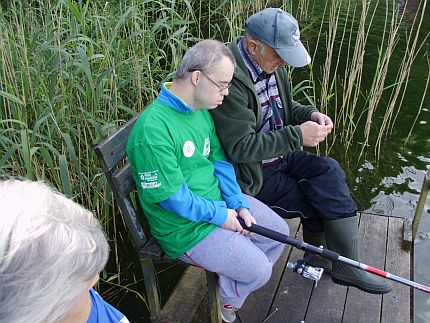 Młody mężczyzna z zespołem Downa siedzi ze starszym mężczyzną na pomoście trzymając wędkę