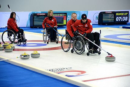 Osoby na wózkach stoją na lodzie, jedna z nich popycha rodzajem kija curlingowy kamień.