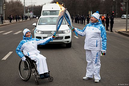 Sztafeta paraolimpijska na ulicy jednego z miast. Dwie osoby, z czego jedna na wózku, przekazując sobie ogień.