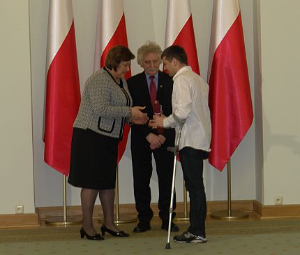 Anna Komorowska i Longin Komołowski wręczają nominację paraolimpijską poruszającemu się o kulach Andrzejowi Szczęsnemu.