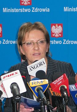 Ewa Kopacz mówi do mikrofonów różnych mediów podczas konferencji, za nią godło z napisem Ministerstwo Zdrowia