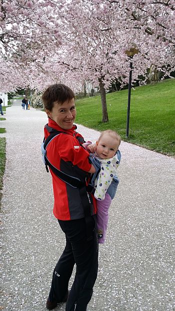 Katarzyna Rogowiec na spacerze z malutką Olimpią w nosidełku. W tle kwitnące drzewa owocowe.