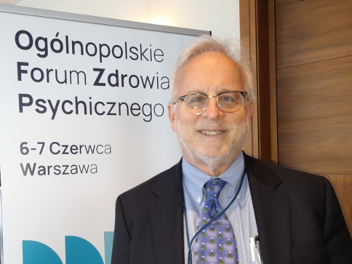 Dr Kenneth Thompson, mężczyzn w średnim wieku, ubrany w garniutur, stoi przy banerze Ogólnopolskiego Forum Zdrowia Psychicznego.